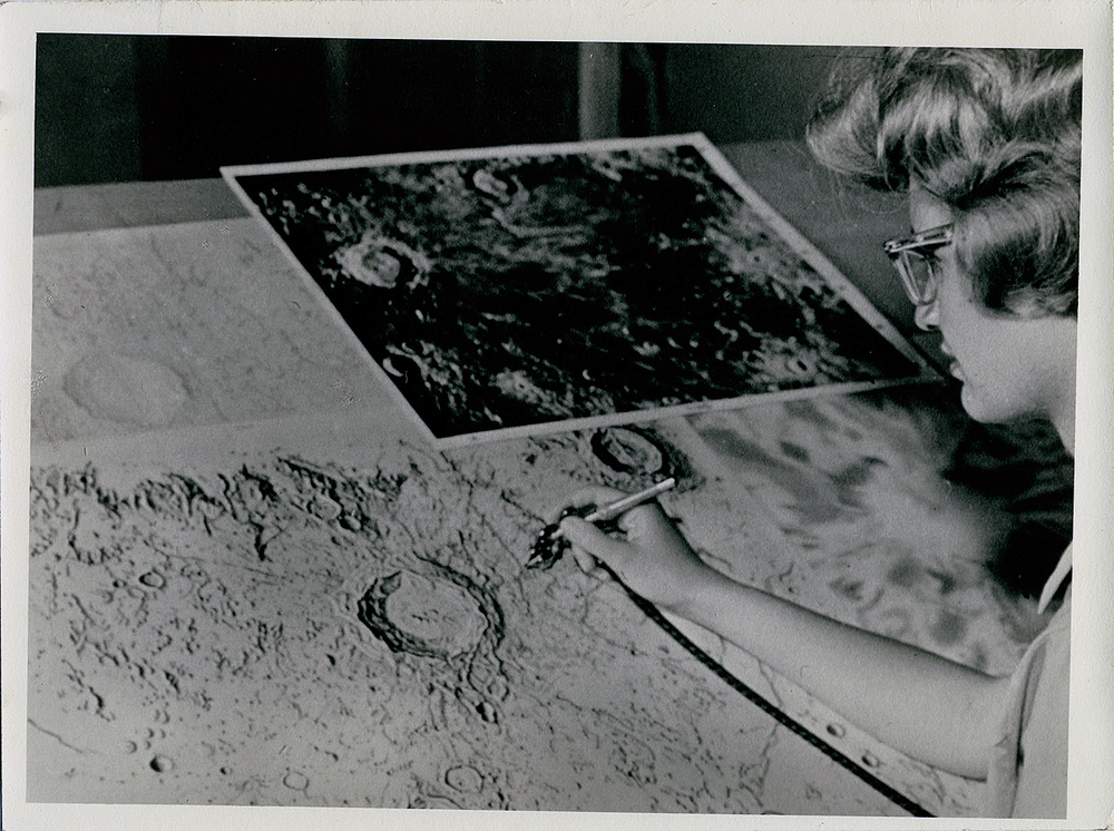 Black-and-white photograph of illustrator Pat Bridges using an airbrush to create detailed maps of the Moon’s surface. She works at a large drafting table covered with lunar topography sketches, referencing a photographic image of the Moon’s craters taken through Lowell Observatory’s Clark Telescope. Bridges leans close to her work, concentrating on capturing the intricate textures and contours of the lunar landscape.