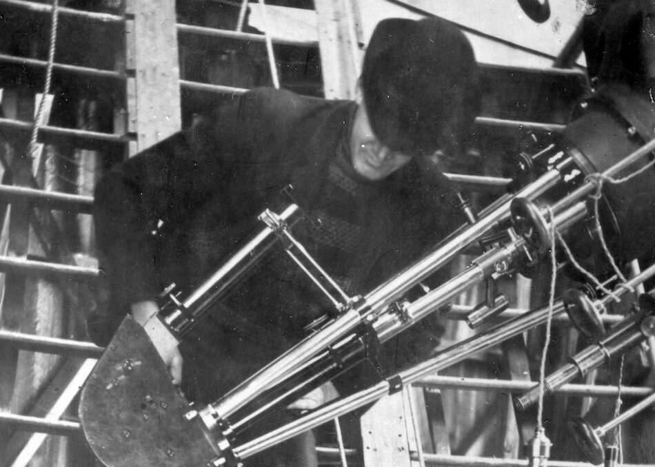 Black-and-white photograph of astronomer V. M. Slipher adjusting a large spectrograph at Lowell Observatory in the early 1900s. Slipher stands on a wooden platform beside the observatory’s telescope mount, surrounded by exposed beams and equipment. A single light bulb hangs nearby, illuminating the scene as he leans over the polished metal instrument, focused on his work.