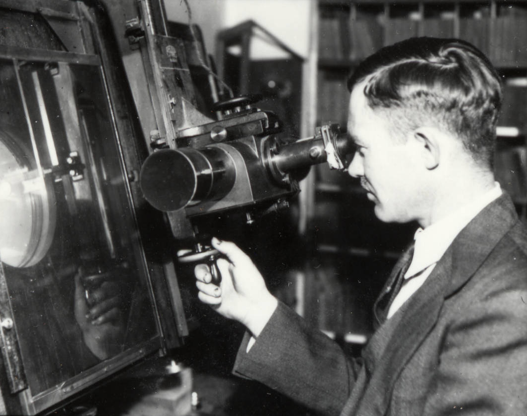 Black-and-white photo of astronomer Clyde Tombaugh examining glass photographic plates using a Zeiss blink comparator at Lowell Observatory. He peers through the eyepiece of the machine, hand on the control lever, comparing star fields that led to the discovery of Pluto in 1930.