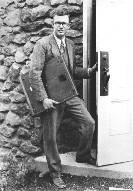 Black-and-white photo of Clyde Tombaugh standing in the doorway of the Pluto Discovery Dome at Lowell Observatory. He holds a large glass photographic plate used for capturing images of the night sky, wearing a suit and tie, symbolizing the era of early astronomical discovery.