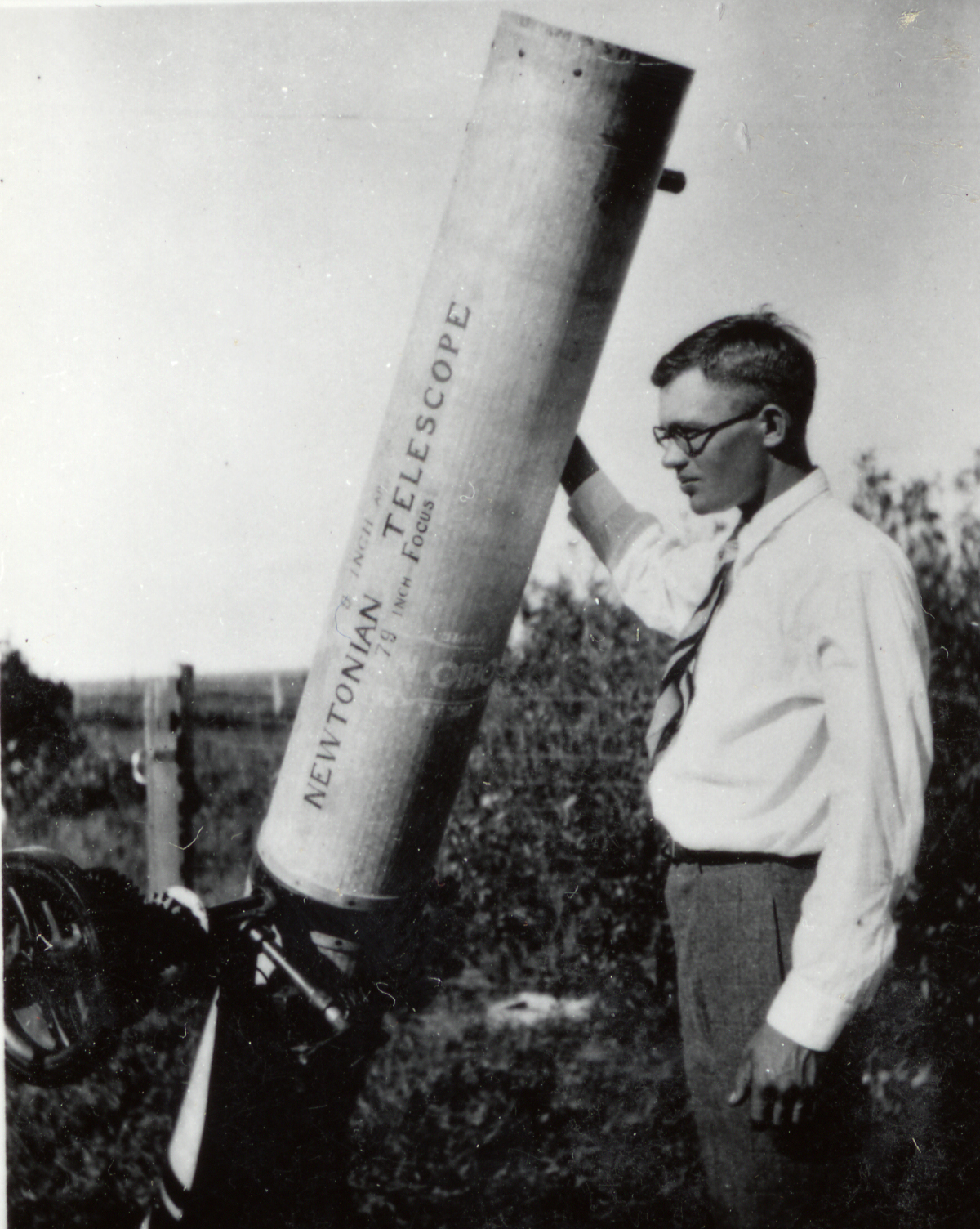 ChatGPT said: Black-and-white photo of Clyde Tombaugh standing outdoors beside his homemade Newtonian telescope. He rests one hand on the long cylindrical telescope tube, wearing a white shirt, tie, and glasses, gazing thoughtfully at the instrument that helped launch his career in astronomy.