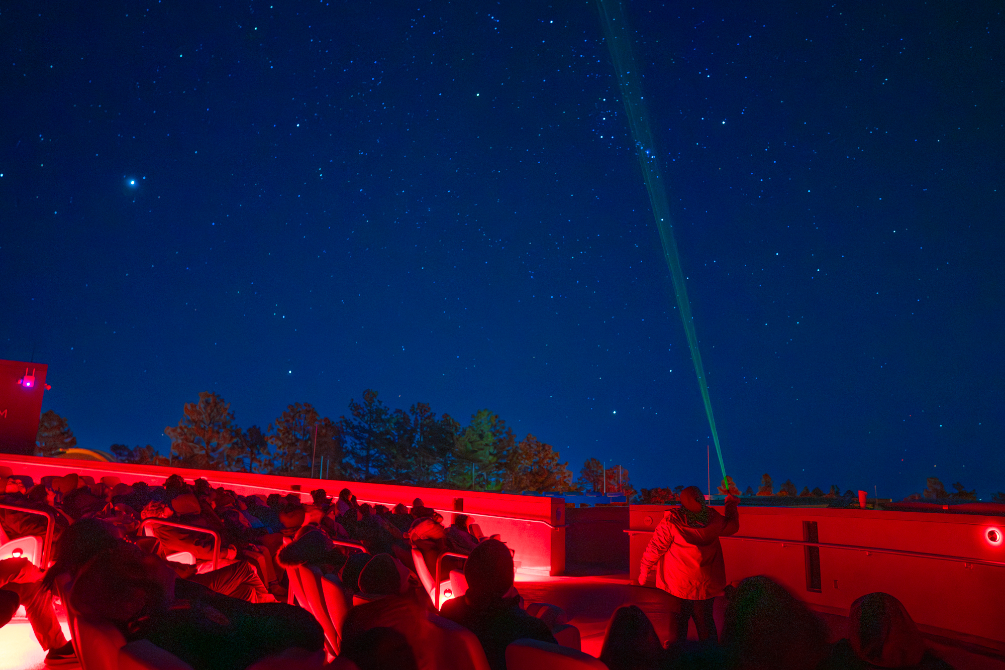 An educator points a green laser pointer toward the star-filled night sky during an outdoor constellation tour at Lowell Observatory, with an audience seated in red-lit chairs among the ponderosa pines of Flagstaff, Arizona.
