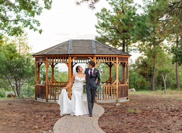 Bride and groom walking in a park gazebo, holding hands, with trees in the background.
