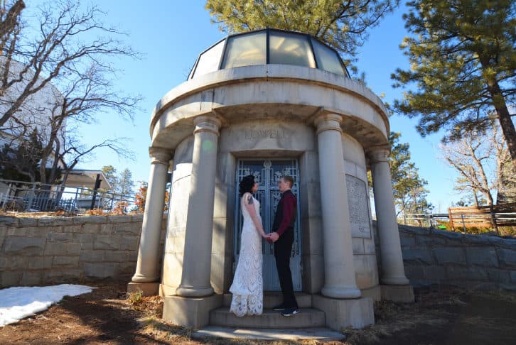 Couple standing in front of Lowell Observatory dome, holding hands, with trees in background.